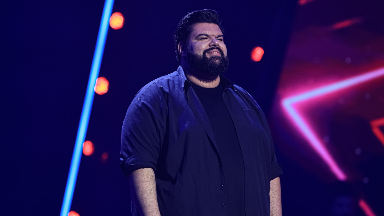 Amadeu Brutus no palco do The Voice Brasil, com barba, camiseta preta e camisa azul aberta, sob iluminação azul e vermelha