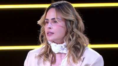 Ana Paula Renault em close, com blazer claro, lenço branco no pescoço e microfone junto ao rosto, diante de um fundo preto com faixas amarelas