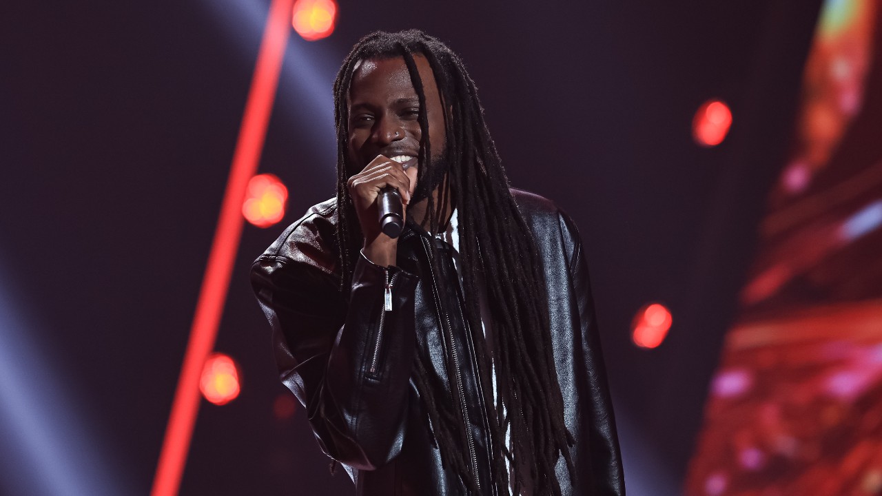 Anderson Mell canta ao microfone com dreadlocks longos e jaqueta preta, diante de luzes vermelhas no palco do The Voice Brasil