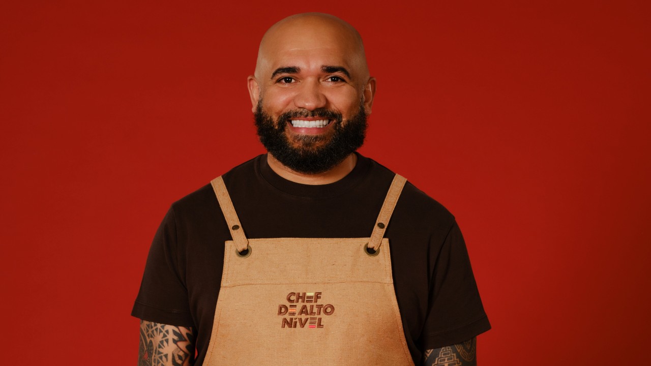 Anderson Sousa sorri com barba, camiseta escura e avental bege de Chef de Alto Nível, diante de um fundo vermelho