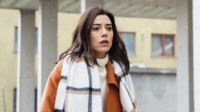 Asya (Cansu Dere) usa casaco marrom e cachecol claro com expressão preocupada em cena da novela Iludida