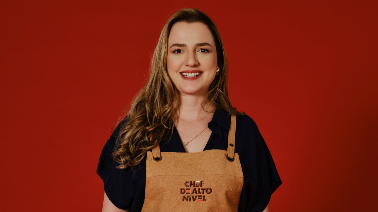Bárbara Cardin sorri com camisa escura e avental bege de Chef de Alto Nível, diante de um fundo vermelho