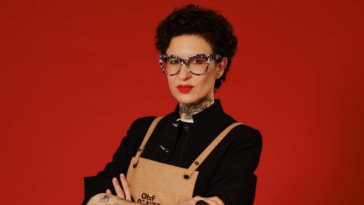 Bárbara Graziela posa de braços cruzados, com óculos estampados, roupa preta e avental bege, diante de um fundo vermelho