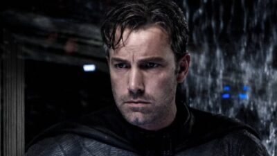 Ben Affleck aparece caracterizado como Batman, com uniforme preto, capa e expressão séria, em uma cena de ambiente escuro