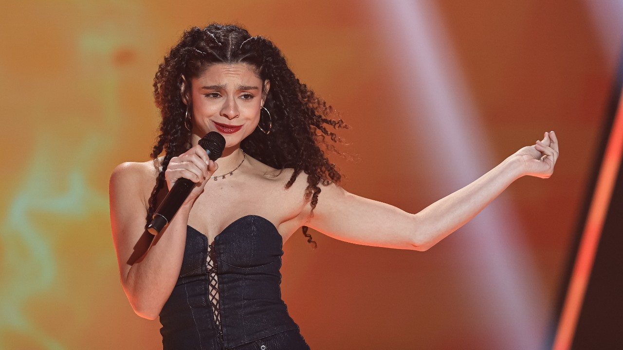Bia Vasconcellos canta ao microfone com roupa escura sem alças e um braço estendido, sob iluminação alaranjada