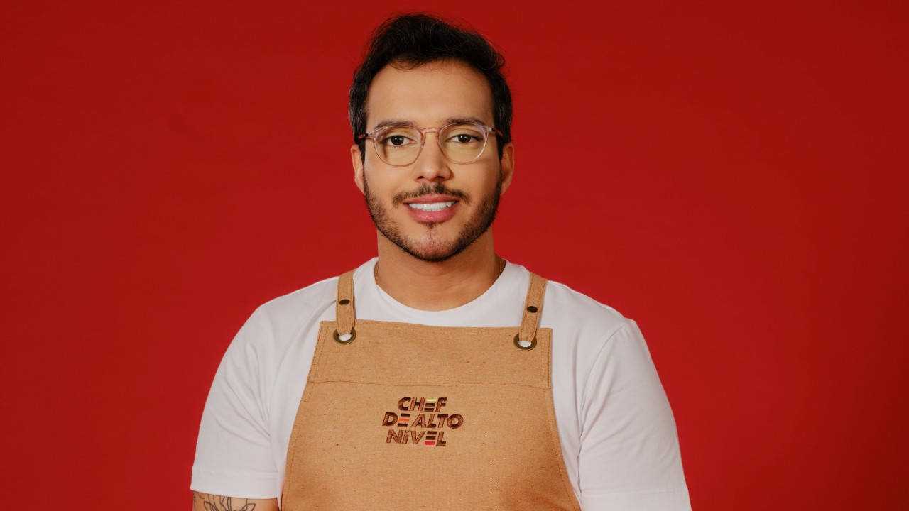 Bruno dos Santos posa com óculos de armação clara, camiseta branca e avental bege de Chef de Alto Nível, diante de um fundo vermelho