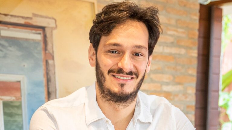 Bruno Luperi aparece sorridente, com camisa branca, barba e cabelos castanhos, em ambiente com paredes de tijolos aparentes e decoração rústica
