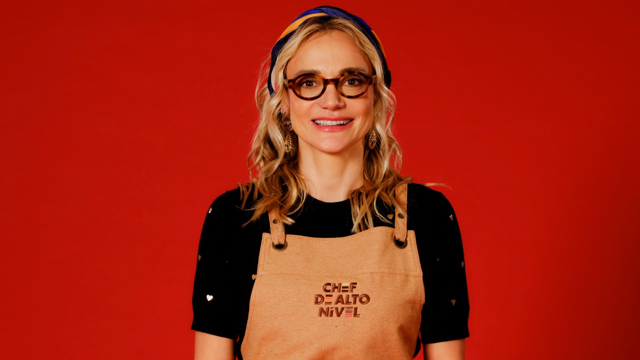 Camila Masullo sorri com óculos marrons, lenço na cabeça, blusa preta e avental bege de Chef de Alto Nível, diante de um fundo vermelho