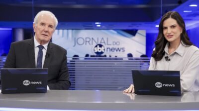 Celso Freitas e Marina Demori aparecem sentados à bancada do Jornal do SBT News, diante de notebooks com a marca do canal, em estúdio com iluminação azul e o logotipo do telejornal ao fundo