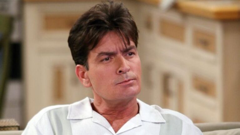 Charlie Sheen em close, com camisa clara de gola aberta e expressão séria