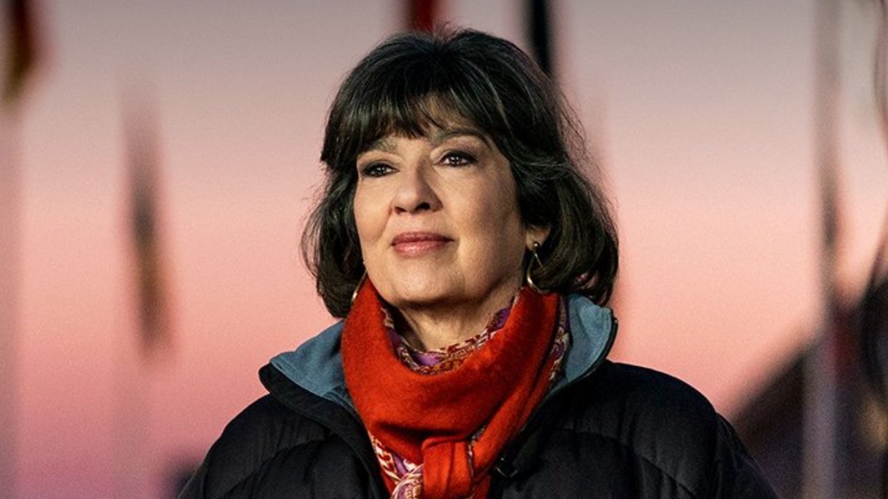 Christiane Amanpour aparece com cabelos escuros e franja, casaco preto e cachecol vermelho, diante de um fundo rosado desfocado