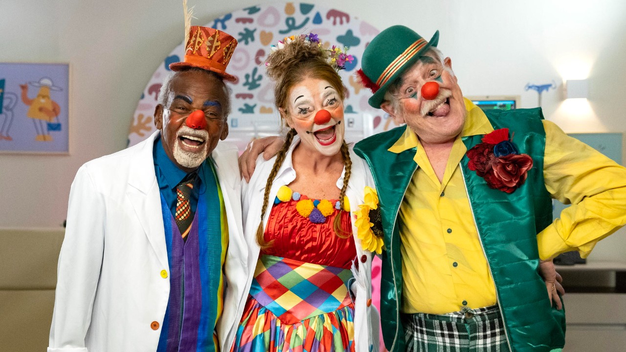 Clemente (Antonio Pitanga), Melinda (Inês Peixoto) e Próspero (José de Abreu) aparecem sorridentes, vestidos de palhaços, com narizes vermelhos e roupas coloridas, em ambiente hospitalar