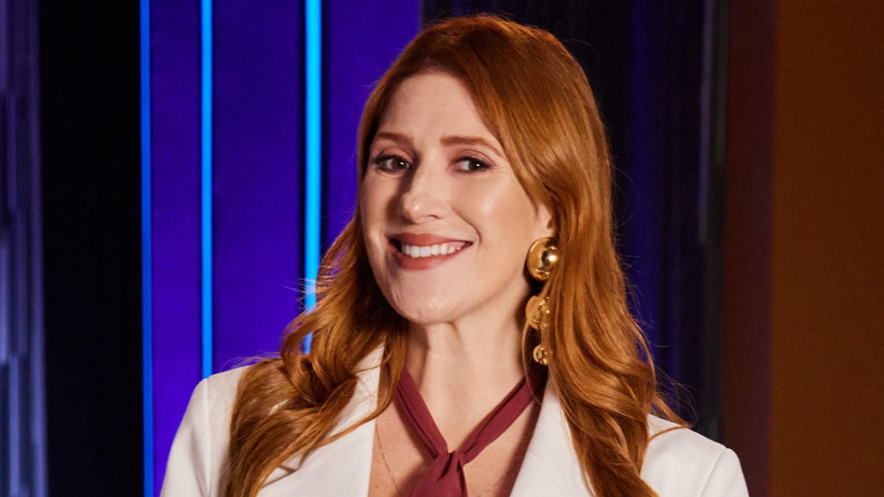Dani Calabresa sorri com cabelos ruivos soltos, blazer branco e brincos dourados diante de cenário iluminado em tons de azul e roxo