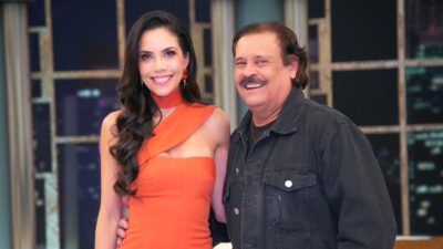 Daniela Albuquerque de vestido laranja e Carlinhos Aguiar de jaqueta preta sorriem juntos em cenário de estúdio