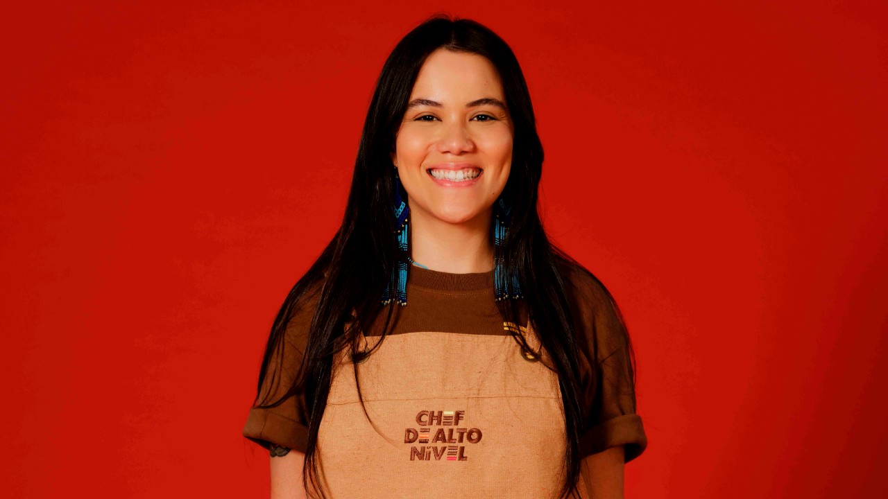 Deborah Pataxó sorri com brincos azuis, camiseta marrom e avental bege de Chef de Alto Nível, diante de um fundo vermelho