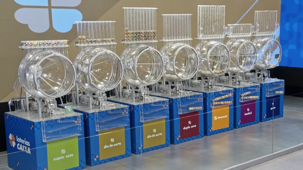 Sete globos transparentes de sorteio alinhados sobre bases azuis das Loterias Caixa, com compartimentos de bolinhas numeradas e placas coloridas que identificam diferentes modalidades