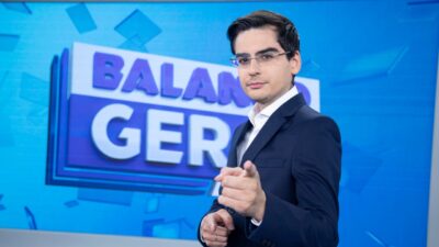 Dudu Camargo de terno escuro e óculos aponta para a câmera diante do telão do Balanço Geral