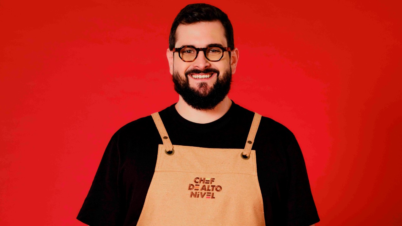 Dudu Teruszkin sorri com barba, óculos pretos, camiseta preta e avental bege de Chef de Alto Nível, diante de um fundo vermelho
