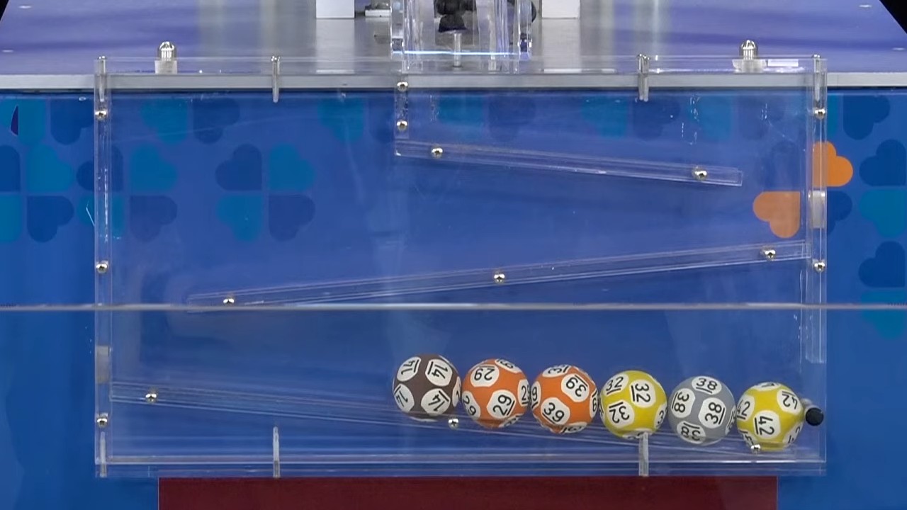 Seis bolinhas coloridas com os números 17, 29, 39, 32, 38 e 42 alinhadas na parte inferior de um equipamento transparente de sorteio, diante de um painel azul