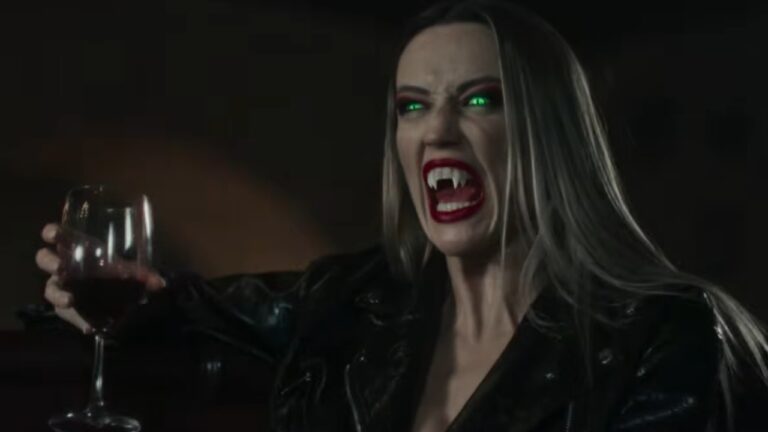 Elizabeth (Mena Suvari) exibe presas de vampira e olhos verdes brilhantes enquanto segura uma taça em Velvet Lounge: Noites de Caça