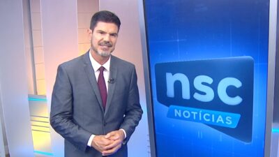 Fabian Londero de terno cinza e gravata vinho apresenta o NSC Notícias ao lado de painel com o logotipo do telejornal