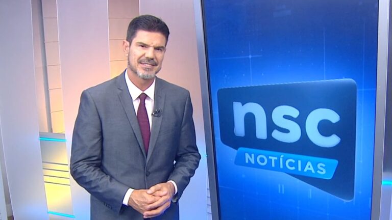 Fabian Londero de terno cinza e gravata vinho apresenta o NSC Notícias ao lado de painel com o logotipo do telejornal