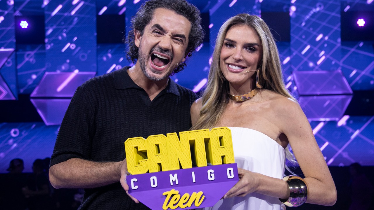 Felipe Andreoli, de camisa preta, e Rafa Brites, de vestido branco, posam sorridentes no palco do Canta Comigo Teen, segurando o logotipo do programa