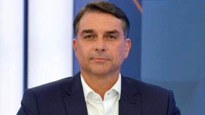 Flávio Bolsonaro aparece de frente, vestindo paletó azul-marinho e camisa branca, diante de um fundo azul