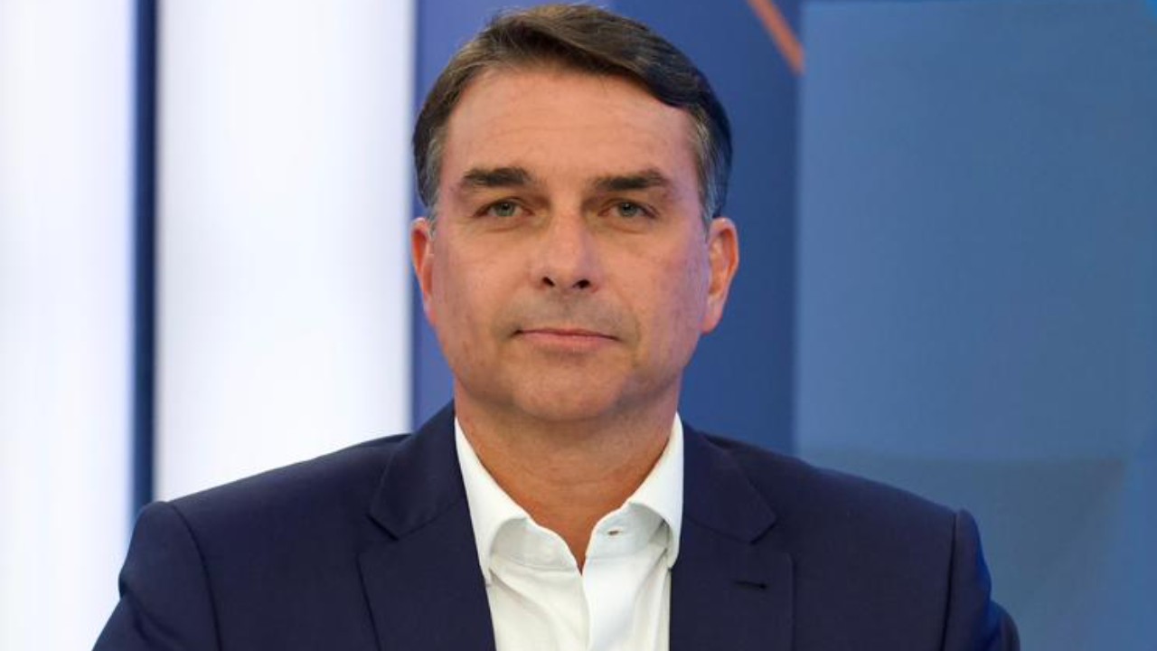 Flávio Bolsonaro aparece de frente, vestindo paletó azul-marinho e camisa branca, diante de um fundo azul