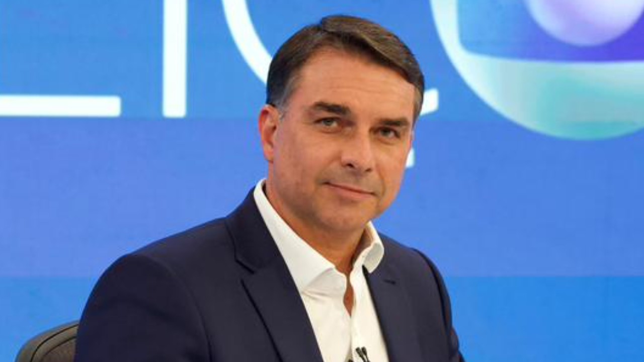 Flávio Bolsonaro aparece em primeiro plano, vestindo paletó azul-marinho e camisa branca, diante de um painel azul em um estúdio de televisão