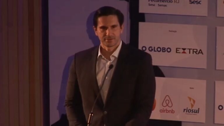 Frederic Kachar, de terno escuro, aparece diante de um microfone durante evento com marcas de empresas ao fundo