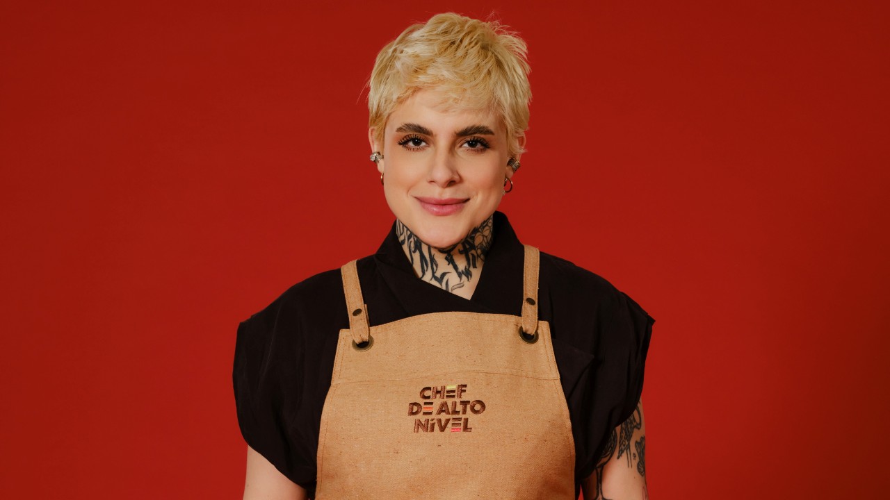 Gabriella Perri posa com cabelo loiro curto, blusa preta e avental bege de Chef de Alto Nível, diante de um fundo vermelho