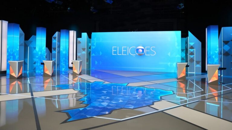 Cenário de debate eleitoral da TV Globo com cinco púlpitos, painéis azuis, mapa do Brasil no chão e Eleições no telão