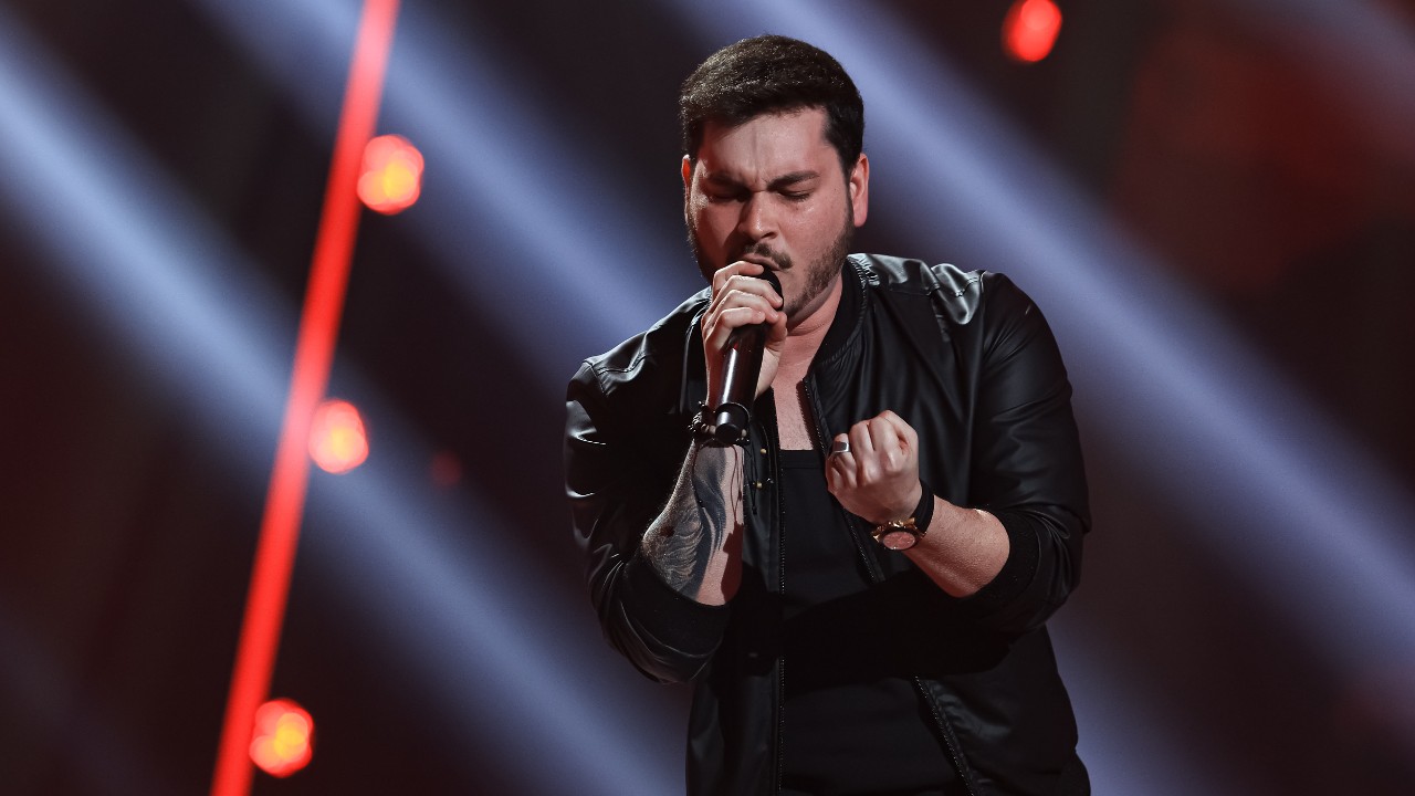 Hudson Araujo canta de olhos fechados ao microfone, com jaqueta preta, diante de luzes vermelhas e brancas