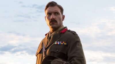 Hughes (Jai Courtney) de uniforme militar e bigode olha para o lado sob céu nublado em Promessas de Guerra