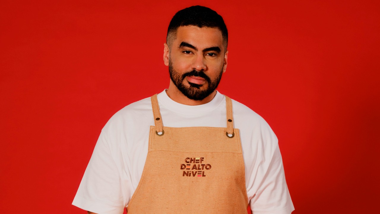 Jamil Máximo posa com barba, camiseta branca e avental bege de Chef de Alto Nível, diante de um fundo vermelho