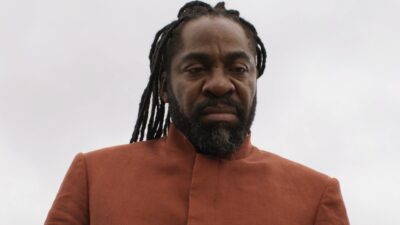 Jendal (Lázaro Ramos) de barba e longos dreadlocks usa traje marrom e encara a câmera sob céu nublado