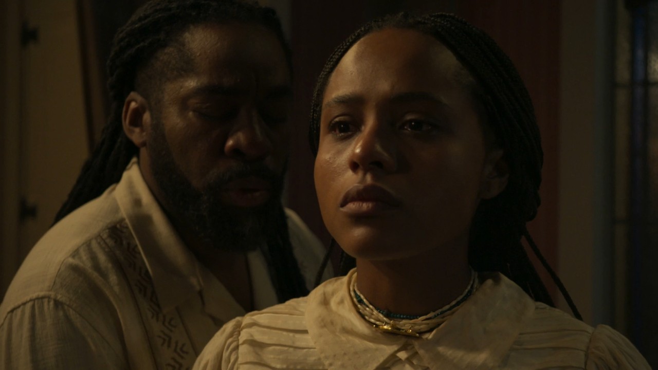 Alika (Duda Santos) com olhar sério e Jendal (Lázaro Ramos) atrás dela em cena da novela A Nobreza do Amor