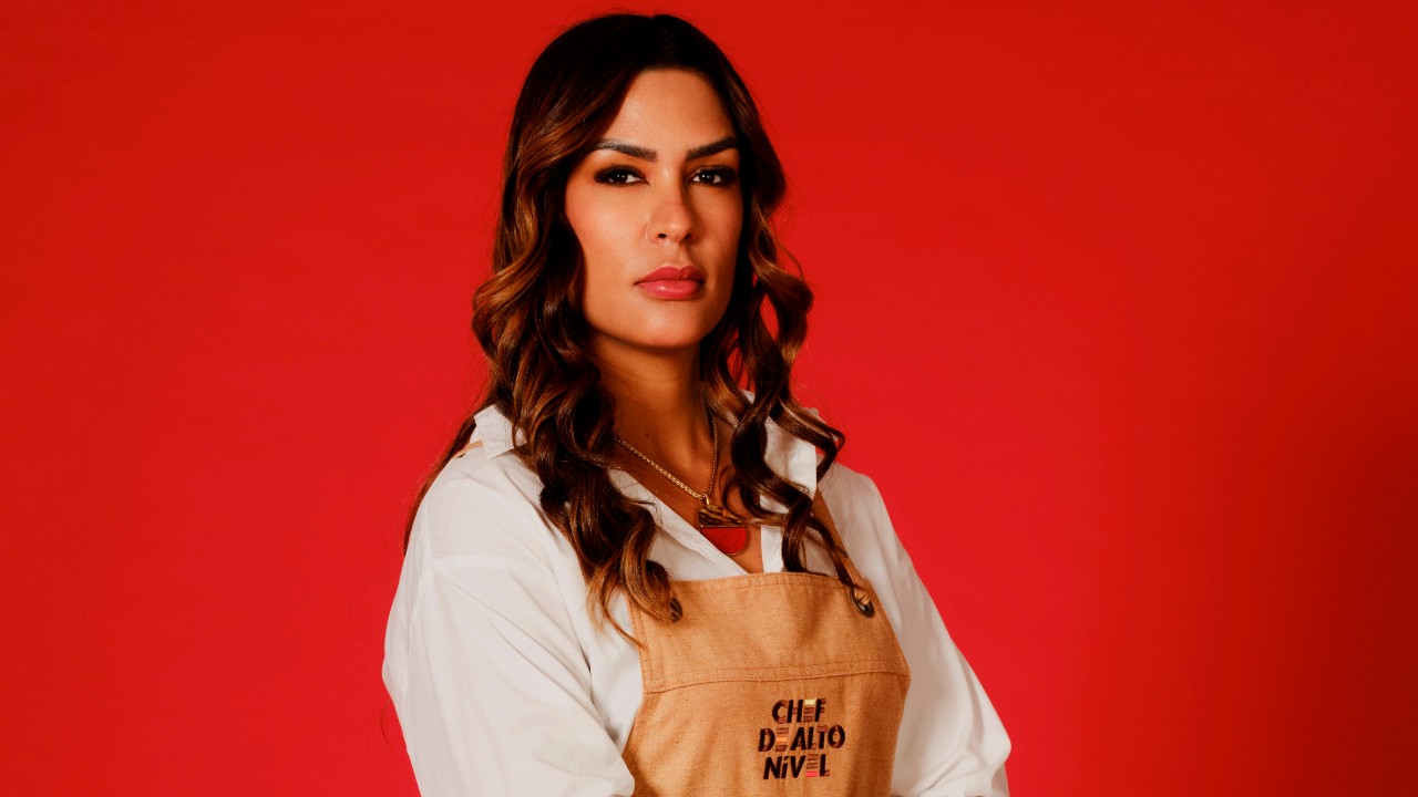 Jéssica Cunha posa com camisa branca e avental bege de Chef de Alto Nível, diante de um fundo vermelho