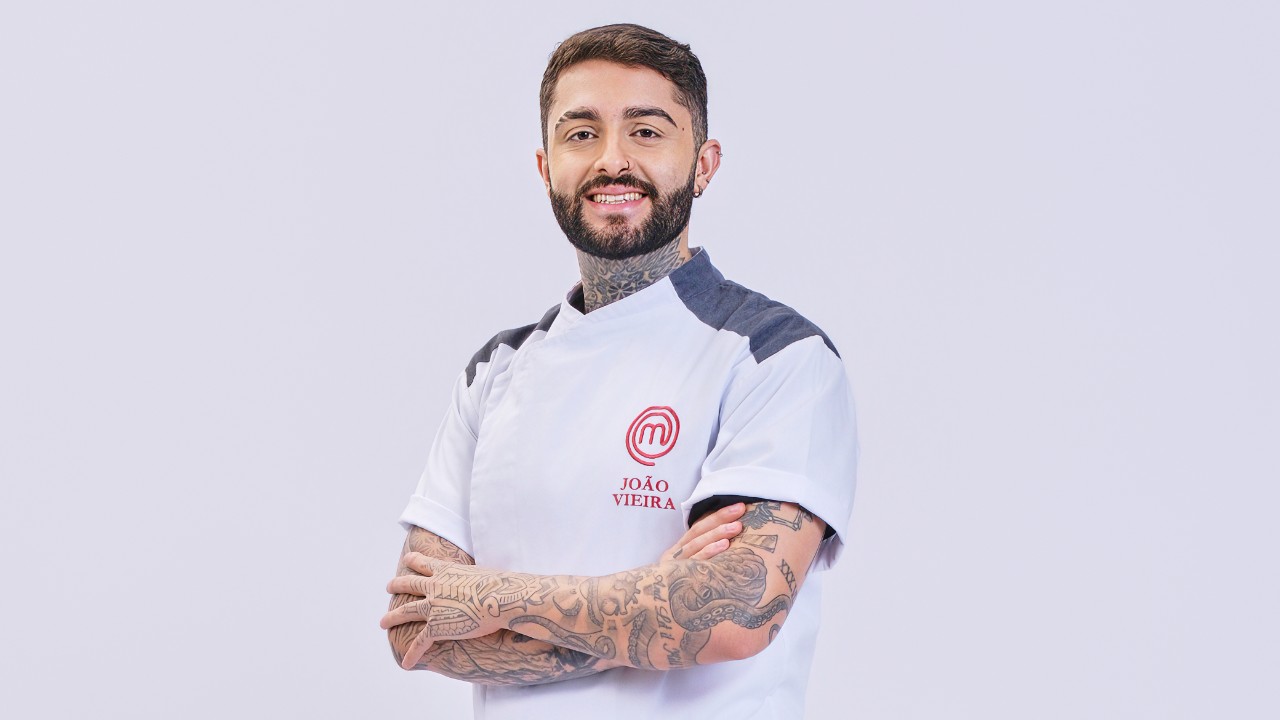 João Vieira posa de braços cruzados com tatuagens e uniforme branco do MasterChef Profissionais