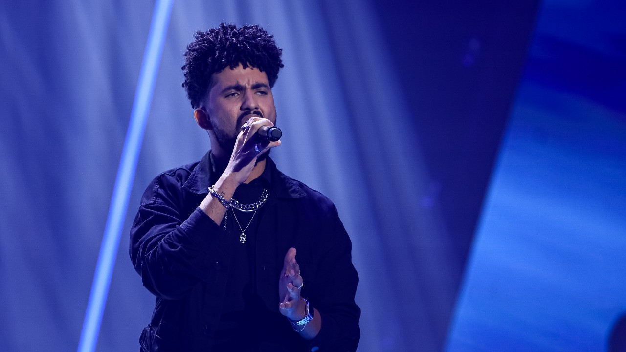 Joe Trindade canta ao microfone com roupa preta, colares e pulseiras, sob feixes de luz azul no palco