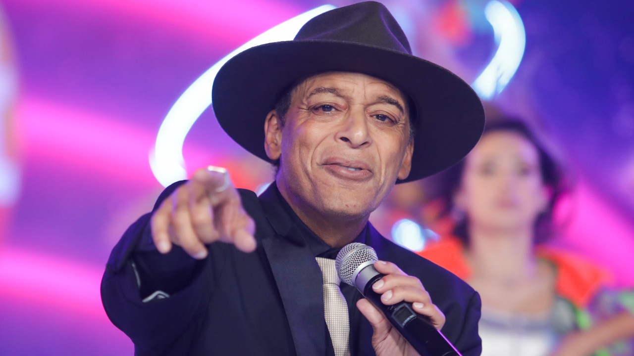 Jon Secada usa chapéu e terno pretos, segura um microfone e aponta para a câmera durante apresentação em palco iluminado