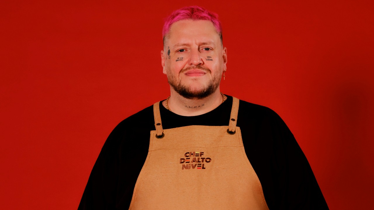 Julio Ritta posa com cabelo rosa, camiseta preta e avental bege de Chef de Alto Nível, diante de um fundo vermelho