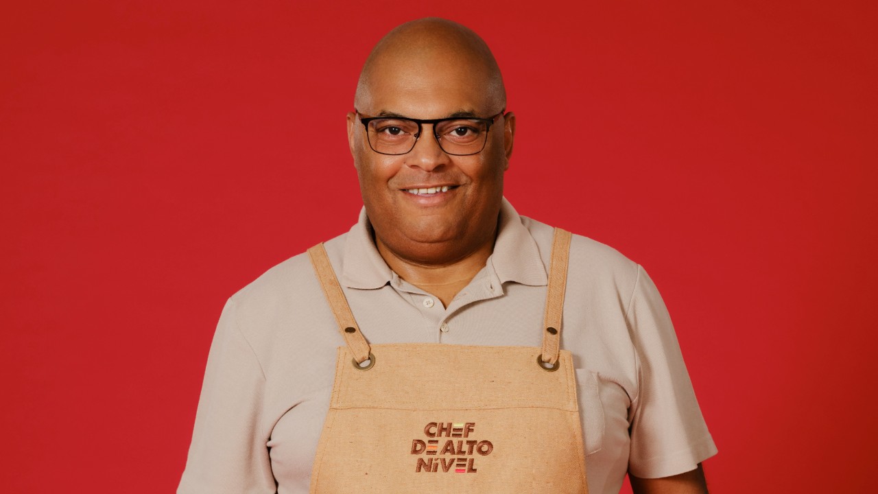 Kiko Silva sorri com óculos pretos, camisa polo clara e avental bege de Chef de Alto Nível, diante de um fundo vermelho