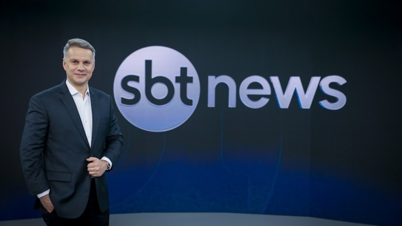 Leandro Magalhães aparece em pé, vestindo terno escuro e camisa branca, diante de um painel azul com o logotipo do SBT News