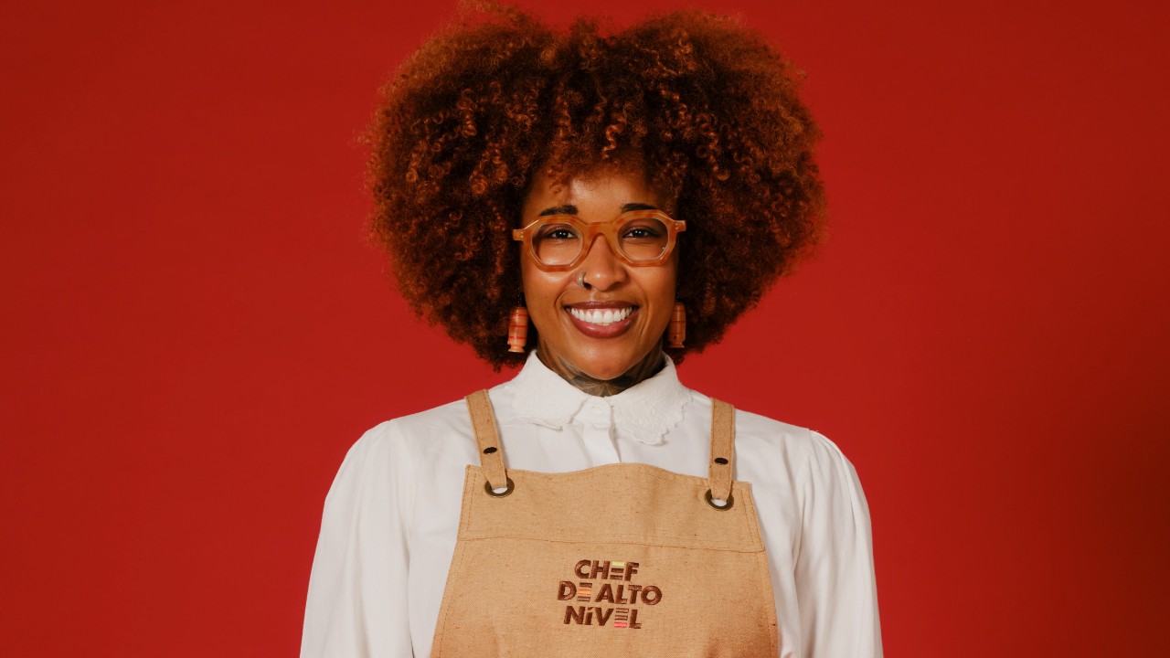 Leila Manhães sorri com cabelos cacheados volumosos, óculos alaranjados, camisa branca e avental bege, diante de um fundo vermelho