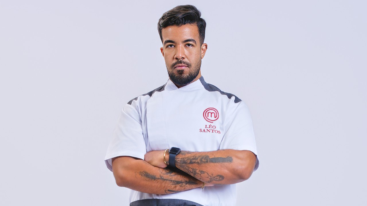 Léo Santos posa de braços cruzados com tatuagens e uniforme branco do MasterChef Profissionais