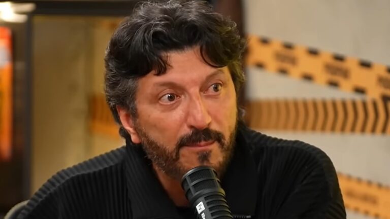 Lito Sousa de roupa preta fala diante de um microfone em estúdio com parede decorada ao fundo