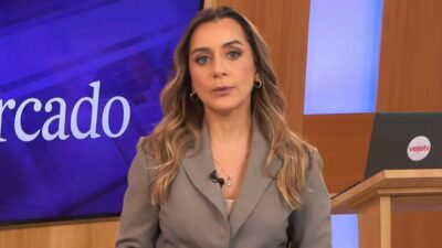 Livia Raick de blazer cinza apresenta telejornal em estúdio, diante de telão azul e púlpito com logotipo da Veja+TV