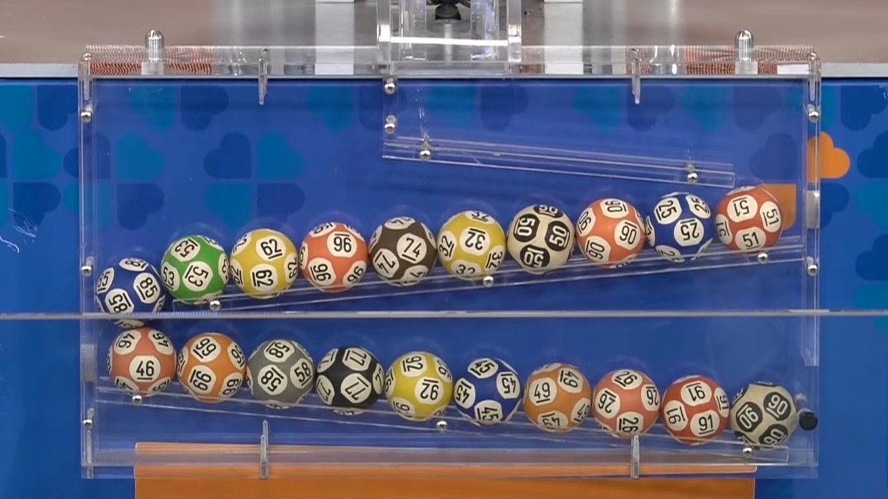 Vinte bolinhas coloridas e numeradas, distribuídas em duas fileiras dentro de um equipamento transparente utilizado no sorteio da Lotomania, com fundo azul e detalhes da identidade visual das Loterias Caixa